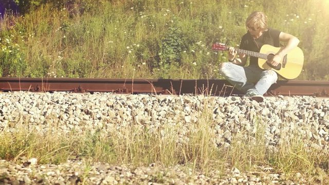 Long-haired guy sits near the railway playing the guitar