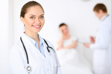 Fototapeta premium Cheerful smiling female doctor on the background with physician and his patient in the bed. High level and quality of medical service concept.