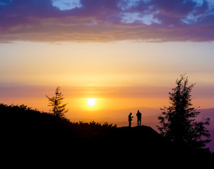 Silhouettes of two people standing on a rock near the tree and looking toward the sun. Sunset in the mountains. Summer in the Ukrainian Carpathians
