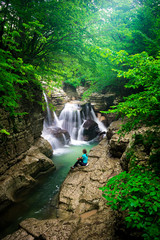 Girl sits in a canyon at the river bank and looks at cascades of water. Trees grows around the river . Sunlight breaks through the green forest. Rest by the river in Georgia