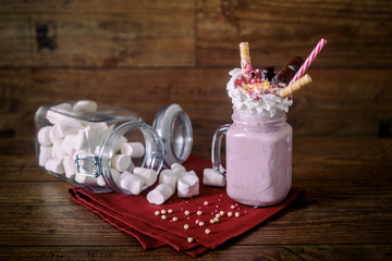 Glass jar of homemade raspberry smoothie cocktail, served with whipped cream, caramel jelly beans and wafer rolls over old dark wood background with jar of marshmallow over background. With copy space