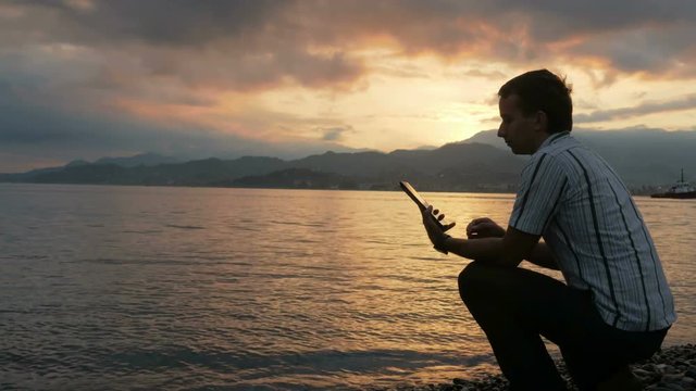 A man in a shirt checks messages on the tablet during the sunrise on the beach of the ocean. Wonderful colors of the sky and the rising of the sun from behind the mountains.