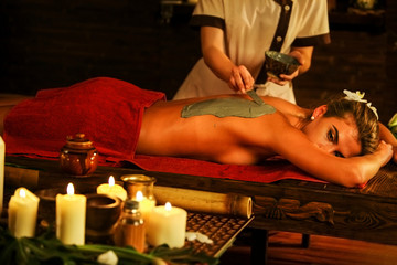 Young woman lying on wooden spa bed. Back massage with clay full body mask in spa salon. Girl on candles background in massage spa salon.