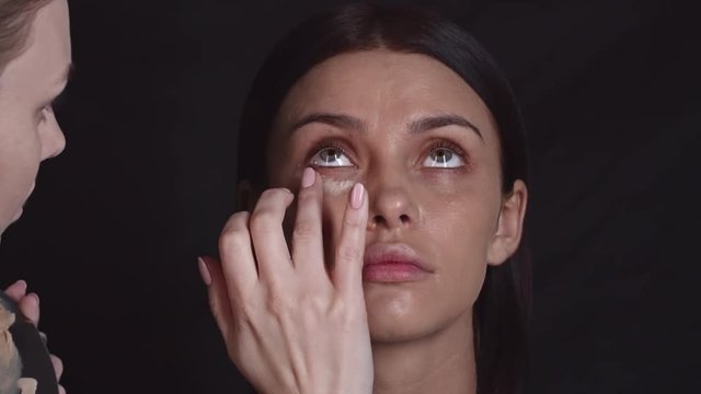 Closeup of professional makeup artist applying concealer under eyes of young woman with fingers and then using brush to blend it