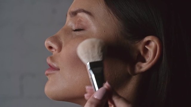 Side view of hands of makeup artist working with brush while applying bronzer to cheeks of woman