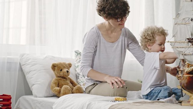 Curly mother and toddler sitting on bed and playing with wooden toys and big sailboat model