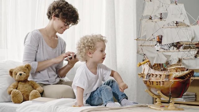 Cute baby boy with curly hair playing with toys and ship model while his mother in glasses chatting with someone via tablet