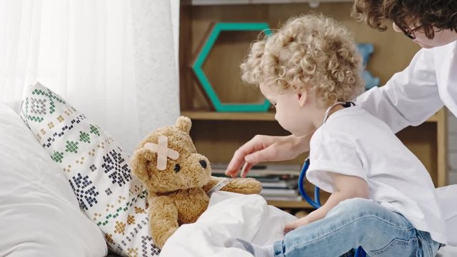 Mother sitting with blond curly toddler playing doctor and stroking sick plush bear 