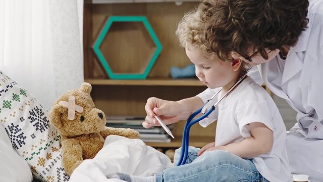 Mother and curly blond toddler playing doctor and measuring temperature for plush bear