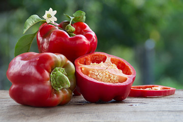 Big red whole and sliced bell pepper on the wooden table. Vegetable on the branch with leaf and flower. Blurred background