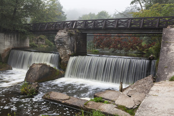 Wooden bridge across the river with waterfall. Foggy morning.