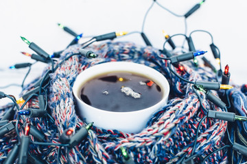 Cup of black tea wrapped in warm woven scarf decorated with fairy lights