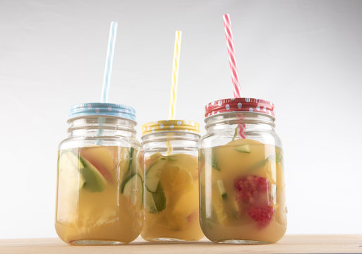 Three glass jars with colourful straws filled with fruit juice and pieces of cut up fruit. Taken on a white background. 