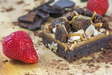 One piece of brownie with chocolate and two strawberries on the light wooden background