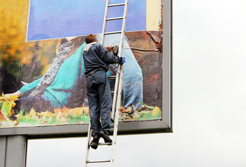 young worker glues a poster with picture of autumn girl on billboard