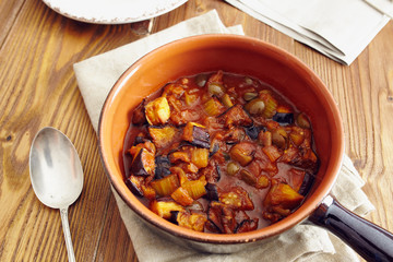 eggplant caponata, traditional sicilian dish. Served in a classic ceramic pan over a napkin on an aged wooden table. Surrounded by a silver spoon and a dish