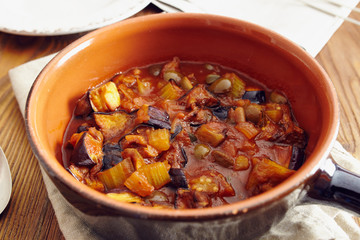 eggplant caponata, traditional sicilian dish. Served in a classic ceramic pan over a napkin on an aged wooden table. Surrounded by a silver spoon and a dish