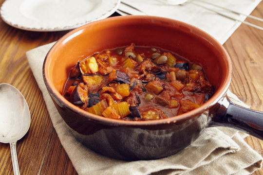 eggplant caponata, traditional sicilian dish. Served in a classic ceramic pan over a napkin on an aged wooden table. Surrounded by a silver spoon and a dish