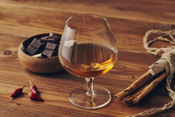 Glass of brandy on a old wooden table with a bowl with chocolate, cinnamon stiks and chili peppers. Warm background.