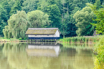 House build on water, straw roof, near lake and green forest.