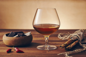 Glass of brandy on a old wooden table with a bowl with chocolate, cinnamon stiks and chili peppers. Warm background.