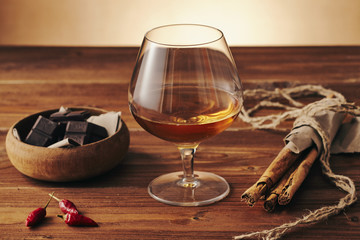 Glass of brandy on a old wooden table with a bowl with chocolate, cinnamon stiks and chili peppers. Warm background.