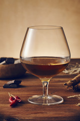 Glass of brandy on a old wooden table with a bowl with chocolate, cinnamon stiks and chili peppers. Warm background.