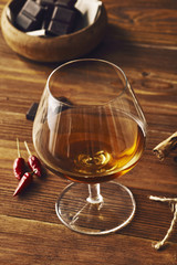 Glass of brandy on a old wooden table with a bowl with chocolate, cinnamon stiks and chili peppers. Warm background.
