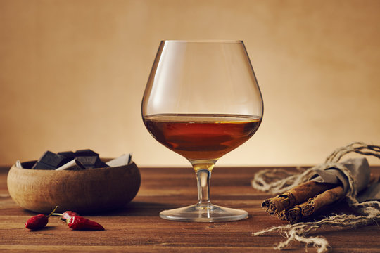 Glass of brandy on a old wooden table with a bowl with chocolate, cinnamon stiks and chili peppers. Warm background.
