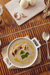 Mushroom cream soup on a table, with bread, garlic, and raw mushrooms on a white chopping board. Salt and Pepper on the table..