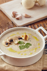 Mushroom cream soup on a table, with bread, garlic, and raw mushrooms on a white chopping board. Salt and Pepper on the table..