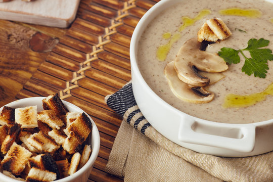 Mushroom cream soup on a table, with bread, garlic, and raw mushrooms on a white chopping board. Salt and Pepper on the table..