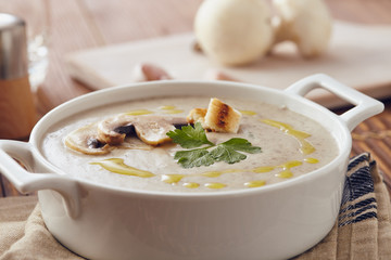 Mushroom cream soup on a table, with bread, garlic, and raw mushrooms on a white chopping board. Salt and Pepper on the table..