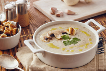 Mushroom cream soup on a table, with bread, garlic, and raw mushrooms on a white chopping board. Salt and Pepper on the table..