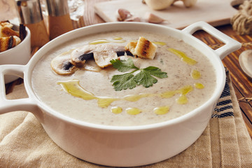 Mushroom cream soup on a table, with bread, garlic, and raw mushrooms on a white chopping board. Salt and Pepper on the table..