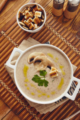 Mushroom cream soup on a table, with bread, garlic, and raw mushrooms on a white chopping board. Salt and Pepper on the table..