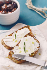 two slice of grilled bread with spreadable cheese and oil on a white chopping board, on a blue wooden table, with olives in a white bowl an rosemary