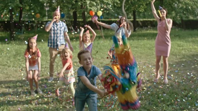 Little boy striking pinata with wooden bat while playing at birthday party in the park and then it falling down in slow motion