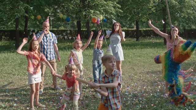 Funny little boy striking pinata with wooden bat while playing at birthday party in the park and then it breaking into two parts in slow motion