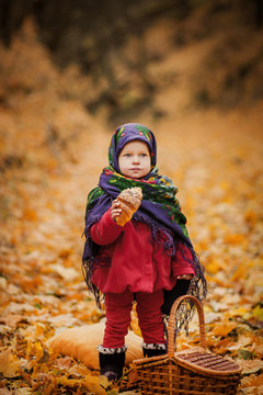 Toddler girl in Ukrainian or Russian folk scarf on head with floral print. Sweet baby eating croissant bun in beautiful autumn forest thickly covered with orange fallen leaves. Portrait of child.