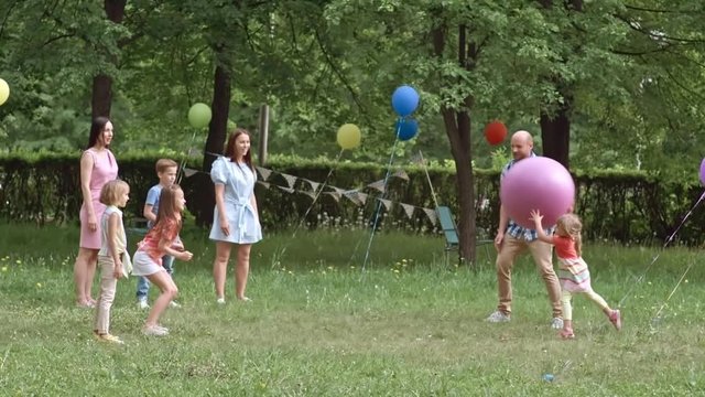 Cute little girl throwing large inflatable ball to her friends while having an outdoor game with parents at b-day party