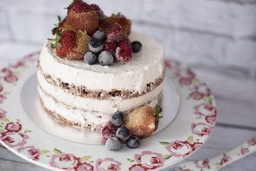 Naked cake with caramelized fruits - strawberries, blueberries, raspberries. Sponge cream cake in floral high plateau, tray. Rustic background
