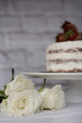 Three white roses. Defocused naked chocolate cake with cream behind. A bright rustic background. Selective focus. Vertical image