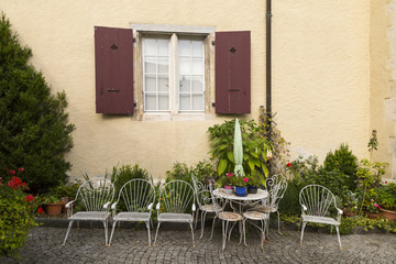 Arrangement mit weissen Gartenmöbeln und Blumen unter einem Fenster mit dunkelroten Fensterläden