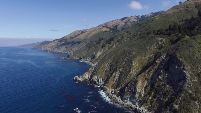 High aerial shot flying by the California Big Sur coast. Deep blue ocean and massive cliffs with waves crashing. Blue skies with sparse clouds.