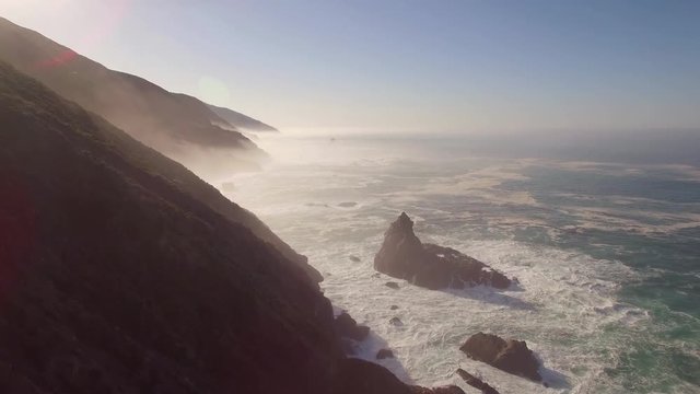 Aerial shot flying by the California Big Sur coast. Steep cliffs and massive rocks in the water. Turquoise blue ocean with waves crashing. Dazzling sun and light mist/fog. 