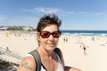 A middle aged woman in sunglasses enjoying visiting Bondi Beach in Sydney, Australia, with short hair, sunglasses, and a tattoo on her shouler.