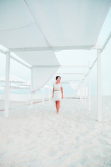 Woman, brunette with short hair in white dress on the beach of ocean, sea near sun shade, tent. Vacation.