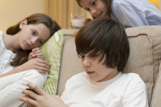 Teenage boy using smartphone while sisters watch in background