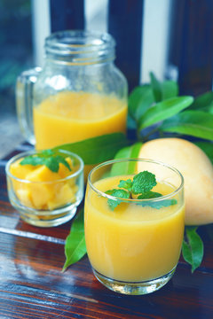 ripe yellow mango smoothie decorated by mint leaf on top with fruit and green leaf on wooden background.